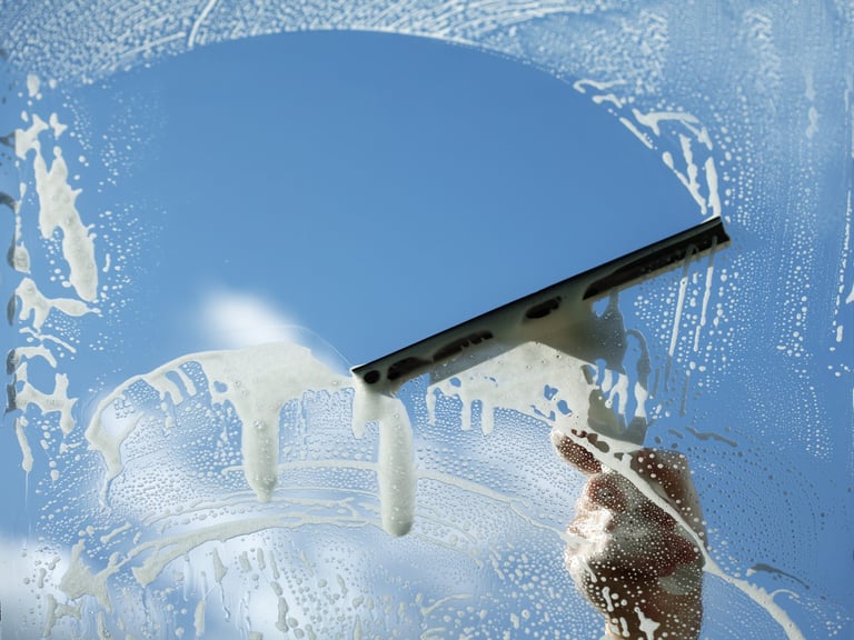 Hand holding a squeegee removing soapy water from a window against a blue sky